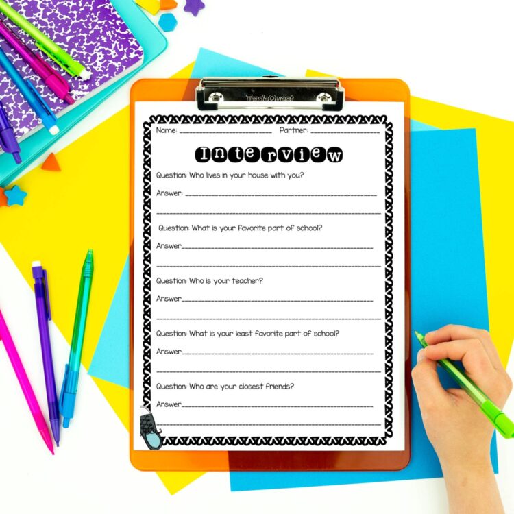 Partner interview page to practice writing in complete sentences while restating the question placed on a clipboard on a desk with a student hand to the right